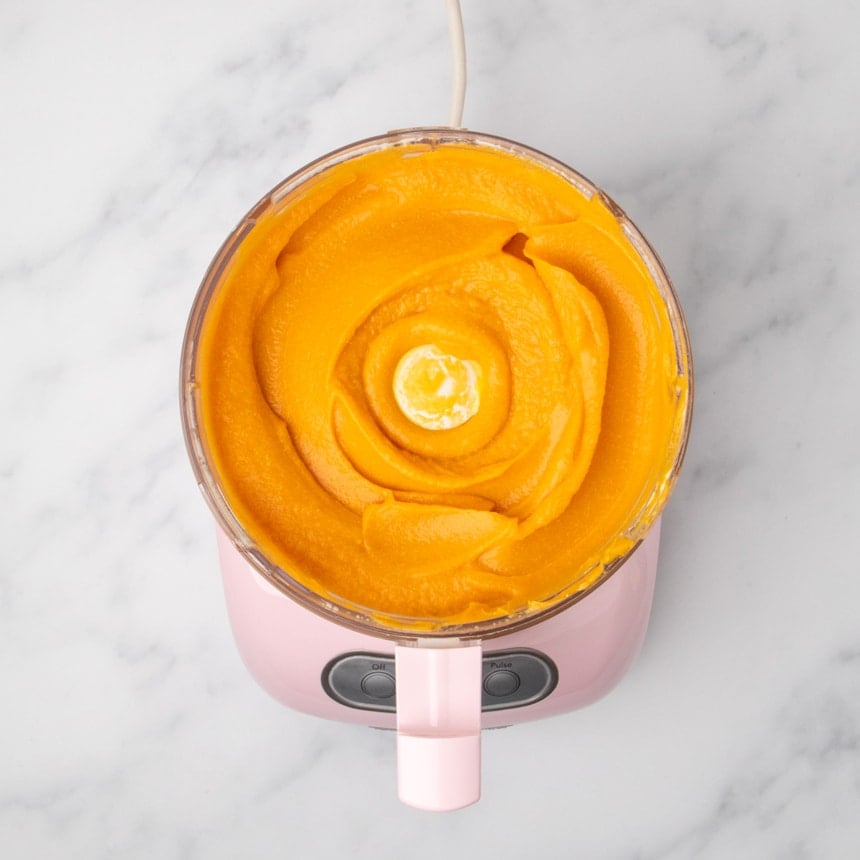 A top view of a food processor filled with smooth orange sweet potato mixture.