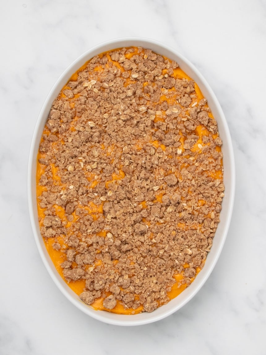 A white oval baking dish filled with orange sweet potato filling topped with an even layer of raw crumb mixture on a white marble surface.