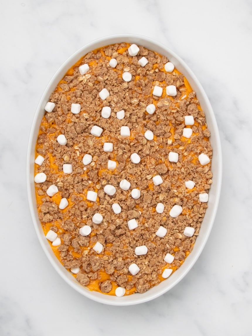 A white oval baking dish filled with orange sweet potato filling topped with an even layer of raw crumb mixture and scattered small white marshmallows, unbaked.