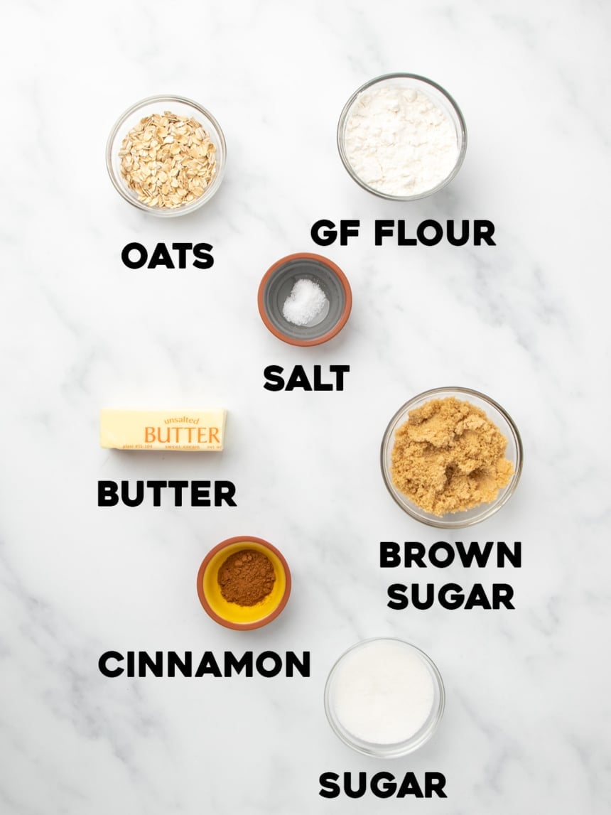 Ingredients labeled with bold text on a white marble surface, including oats, gluten free flour, salt, butter, brown sugar, cinnamon, and white sugar.