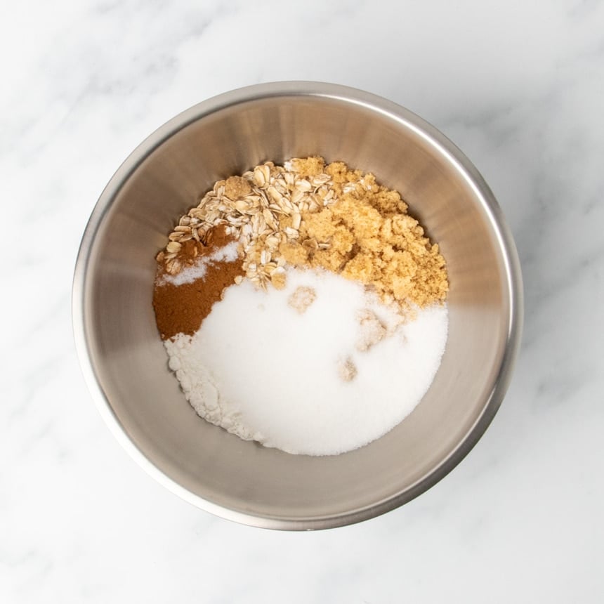 A metal mixing bowl containing dry ingredients including oats, brown sugar, white sugar, gluten free flour, and cinnamon on a white marble surface.
