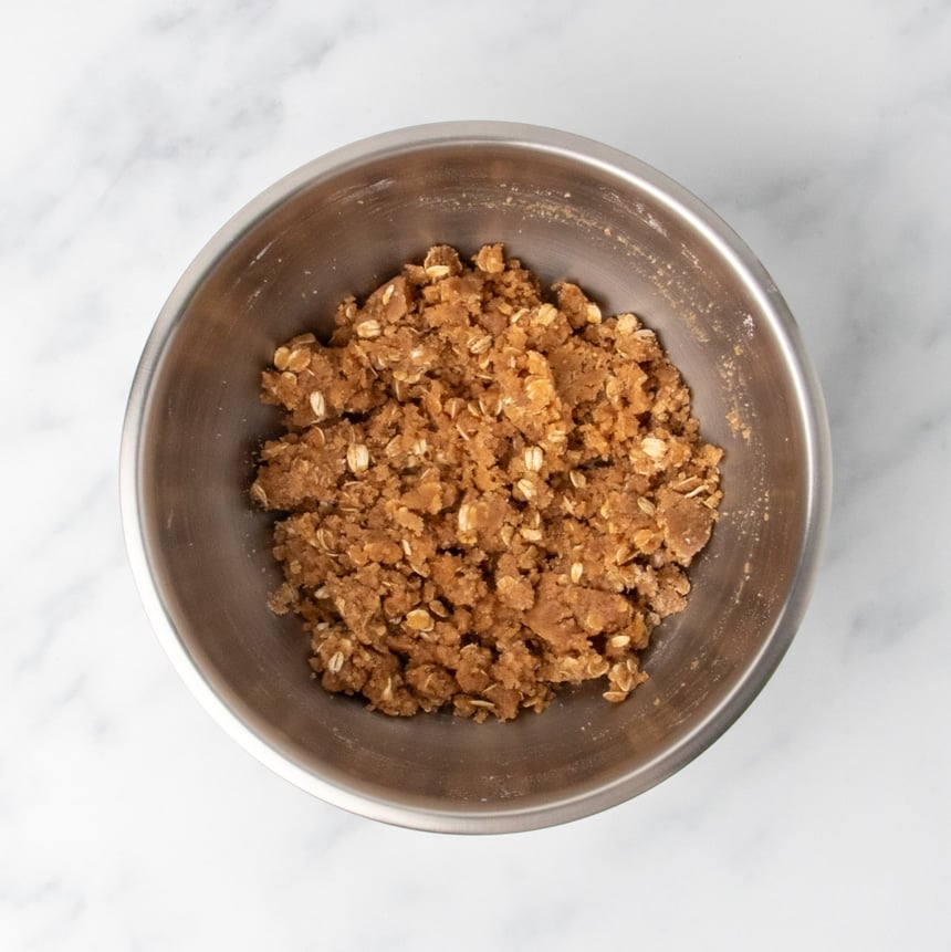 A metal mixing bowl filled with a dark brown, clumpy mixed oat and sugar topping mixture.