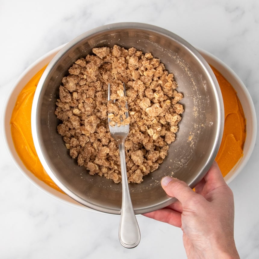 A hand holding a metal bowl with a fork in a crumbly oat topping mixture above a dish filled with orange sweet potato filling.