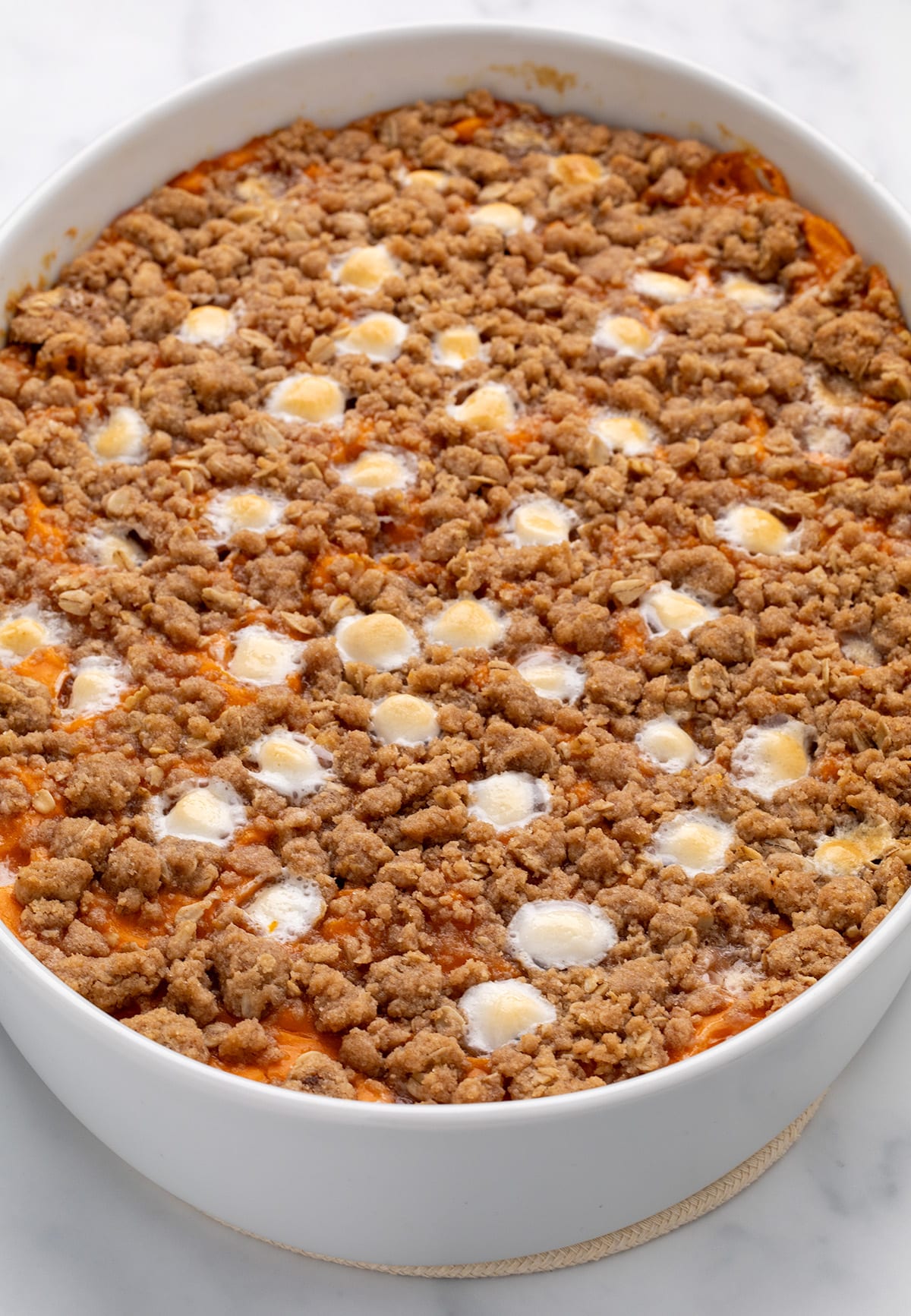 A close-up of a baked gluten free sweet potato casserole in a white oval dish, showing a browned crumb topping with melted white marshmallows on the surface.