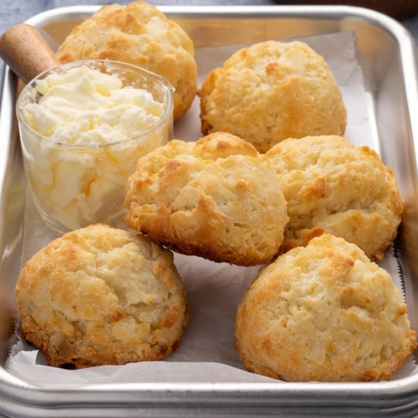 closeup of 6 small gluten free drop biscuits on a small metal tray with a glass container filled with whipped butter