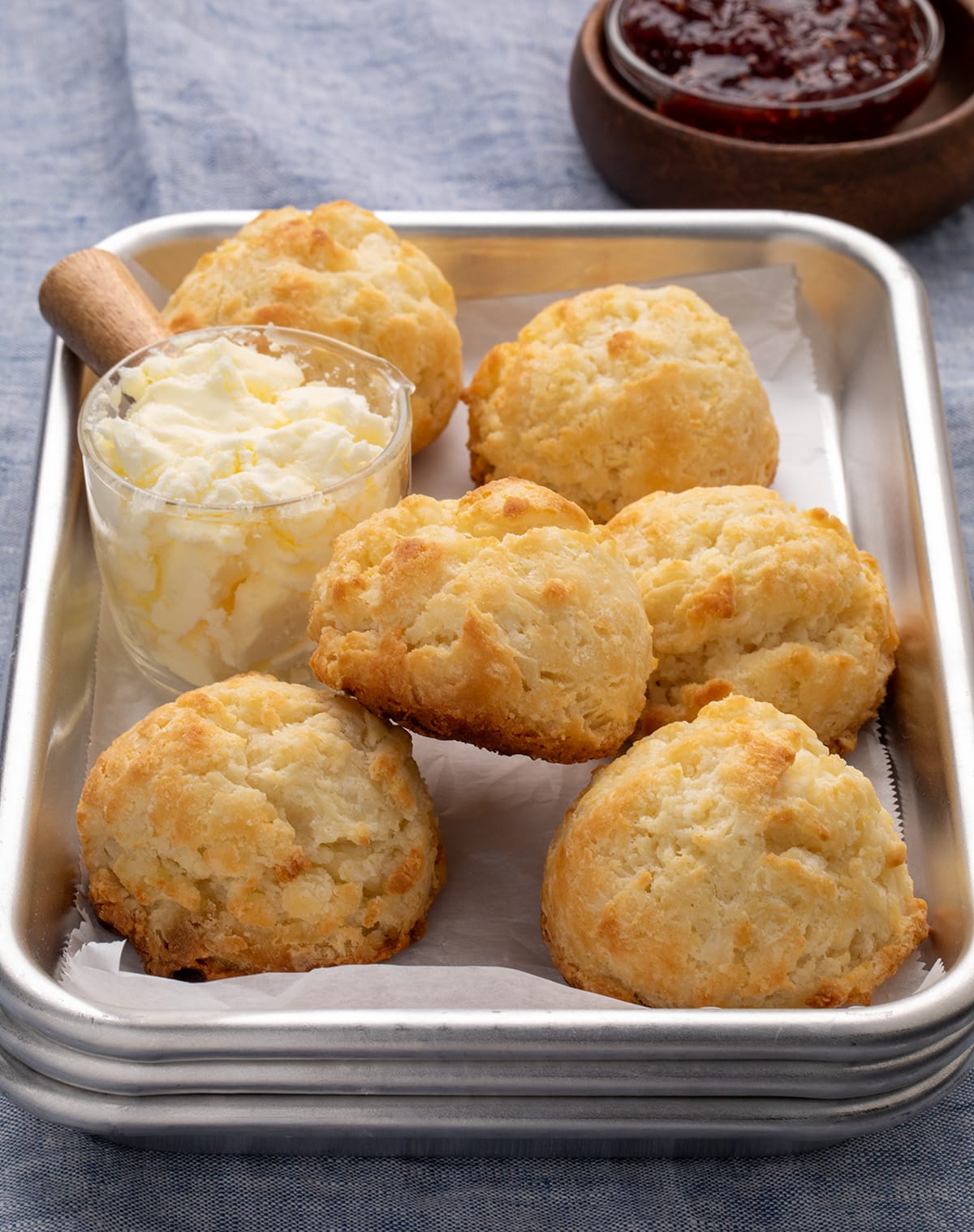 small metal 1/8 sheet pan tray lined in white paper with 6 golden brown drop biscuits on top with a a small glass bowl of pale yellow butter on a blue cloth
