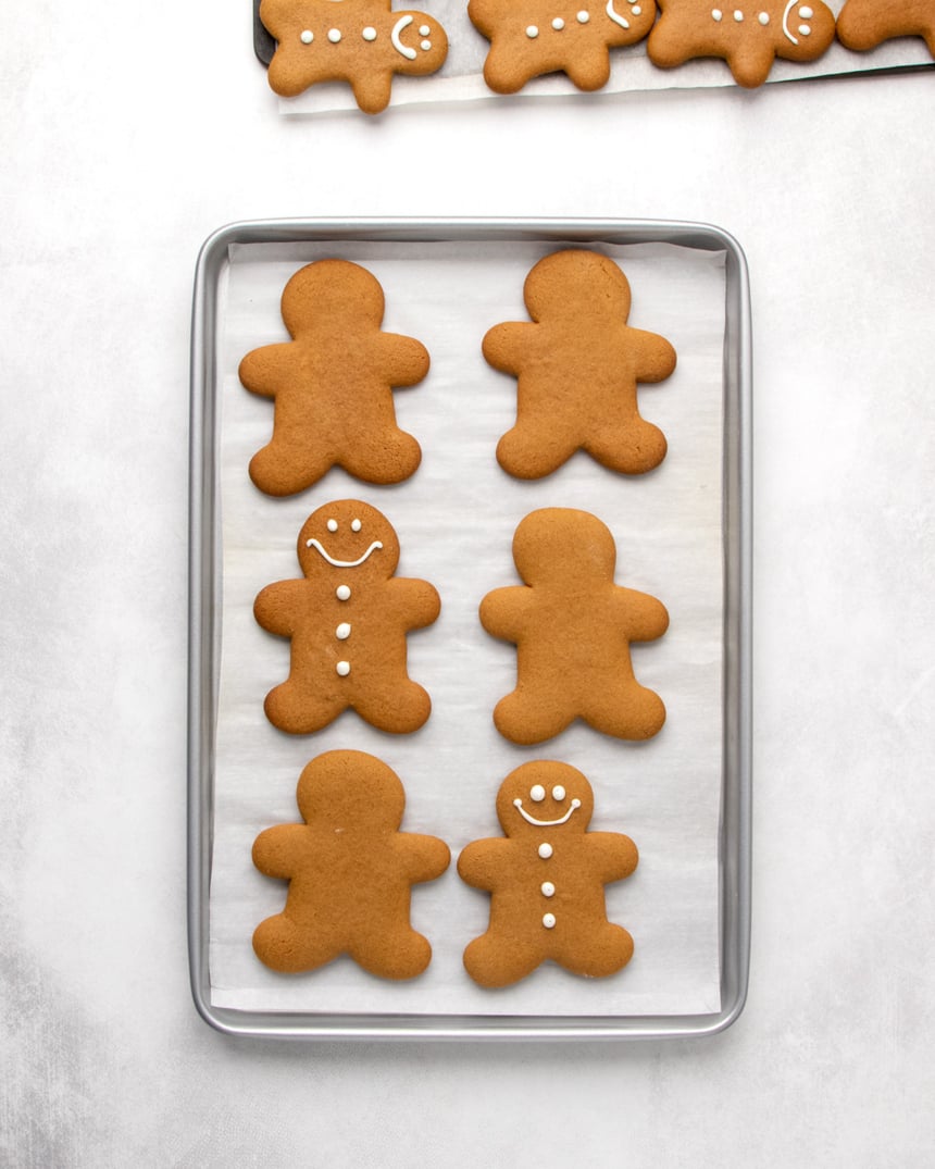 A metal baking sheet lined with white parchment paper holding six large brown gingerbread men-shaped cookies, two of which have simple white icing faces and buttons. Additional decorated cookies are partially visible on a baking sheet at the top edge of the image.