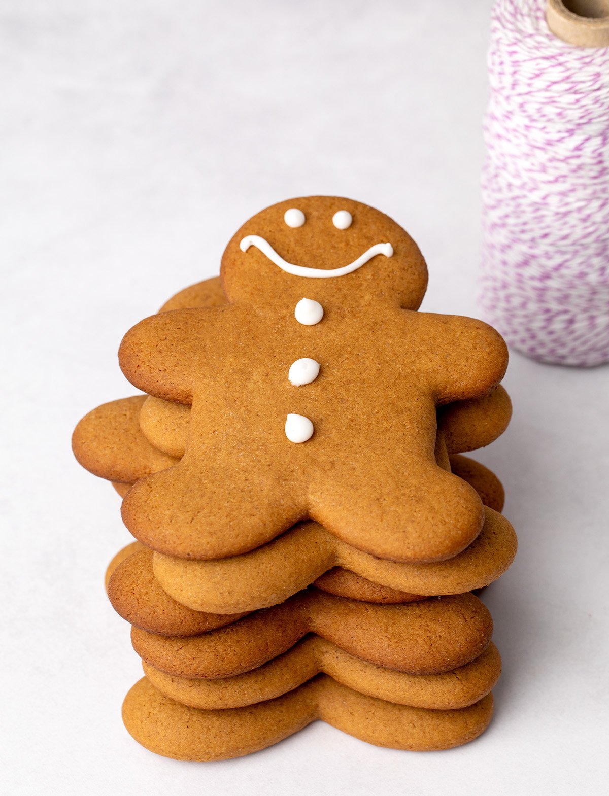 A stack of 6 large gingerbread men shaped cookies with one iced cookie on top decorated with a simple white icing face and buttons. A spool of purple-and-white twine is in the background.