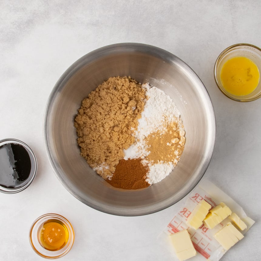 A metal mixing bowl containing brown sugar, gluten free flour, cinnamon, ginger, salt, and baking soda. Surrounding the bowl are small containers of molasses, honey, beaten egg, and cubed butter on its wrapper.