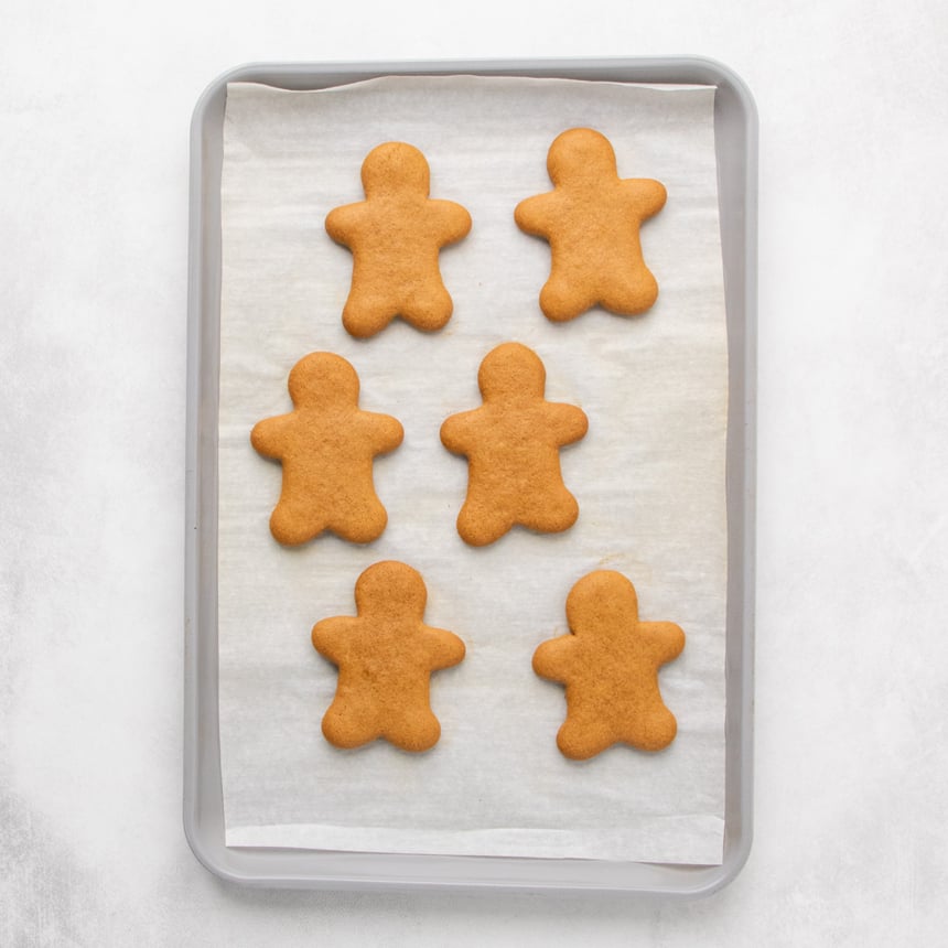 A parchment-lined baking sheet with six brown baked gingerbread-person cookies arranged in two columns.