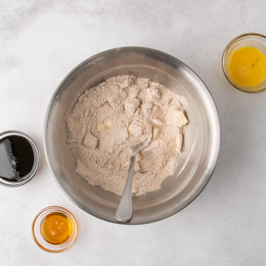 A metal mixing bowl filled with the dry ingredients mixed together, with visible pieces of butter. A spoon rests inside the bowl. Containers of molasses, honey, and beaten egg sit nearby.
