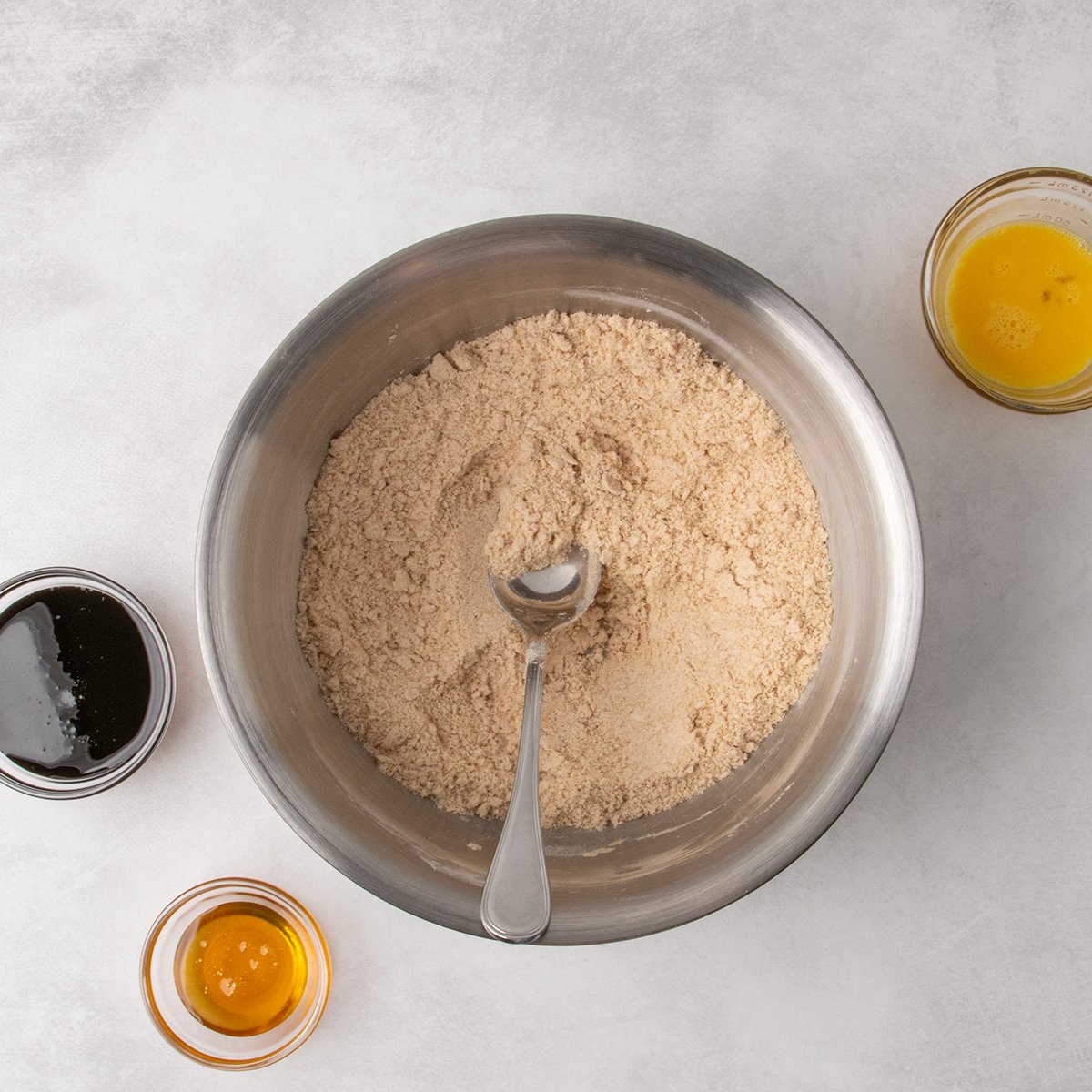 A metal mixing bowl filled with a uniform crumbly mixture of dry ingredients with softened butter mixed in until the mixture resembles coarse sand. A spoon rests inside the bowl. Containers of molasses, honey, and beaten egg are placed around the bowl.