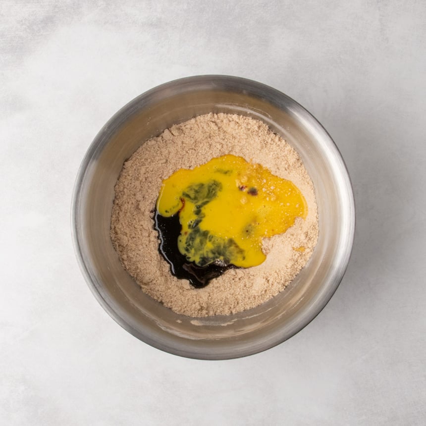 A metal mixing bowl with the crumbly mixture, topped with poured molasses, honey, vanilla, and beaten egg.