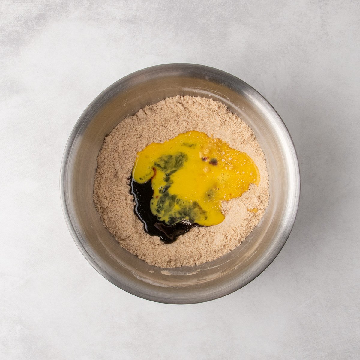 A metal mixing bowl with the crumbly mixture, topped with poured molasses, honey, vanilla, and beaten egg.
