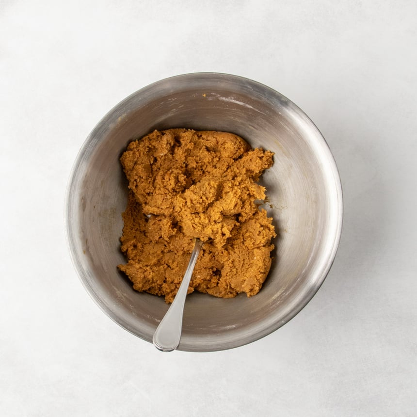 A metal mixing bowl containing fully mixed thick, brown gingerbread cookie dough with a spoon sticking out of it.
