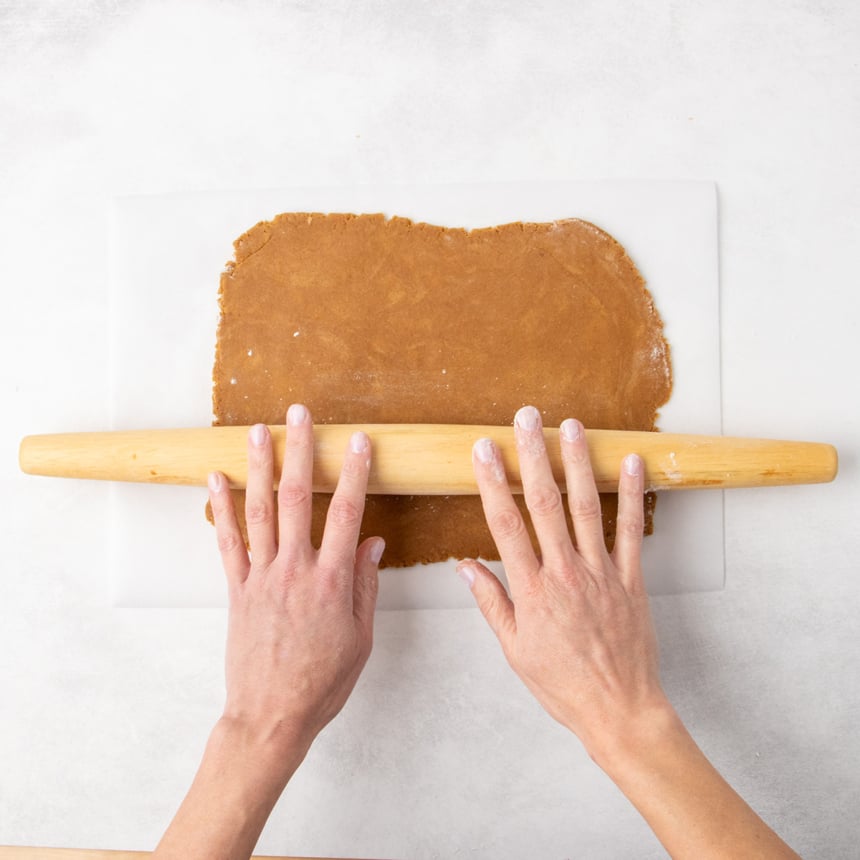 Two hands using a rolling pin to roll out raw brown cookie dough on parchment paper.
