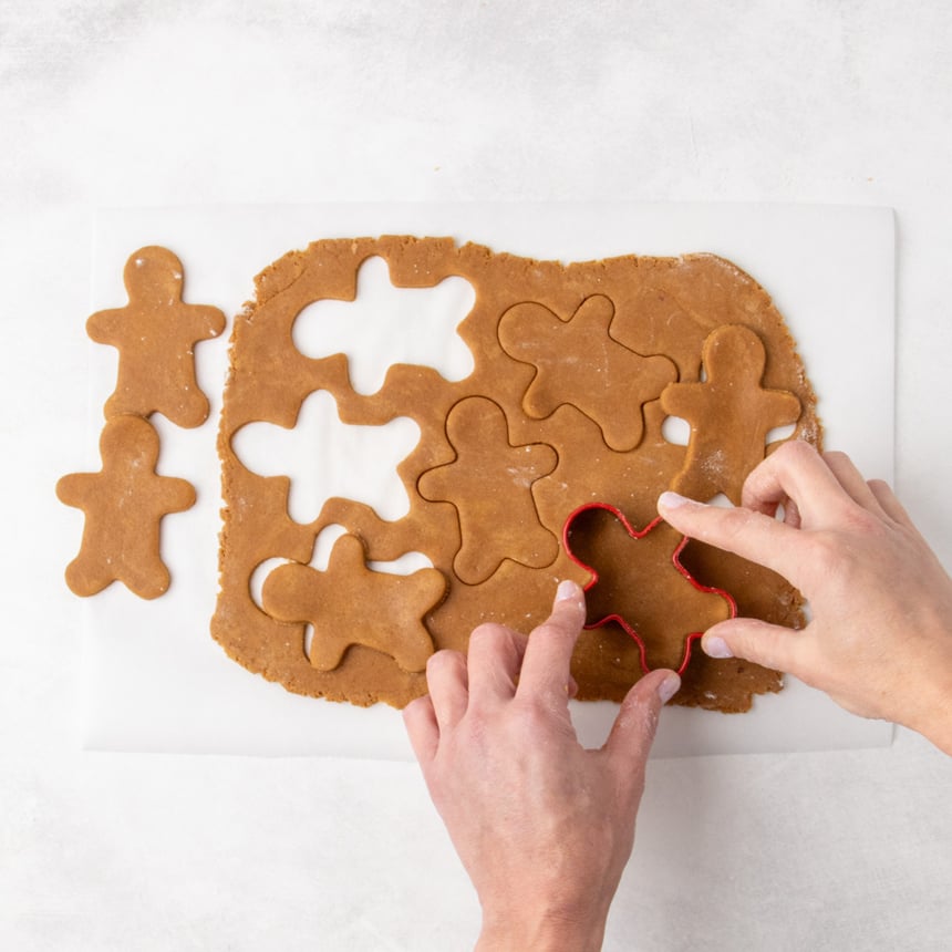 Two hands pressing a red gingerbread-person cookie cutter into rolled raw brown cookie dough. Several cut-out shapes and remaining dough surround the cutter.
