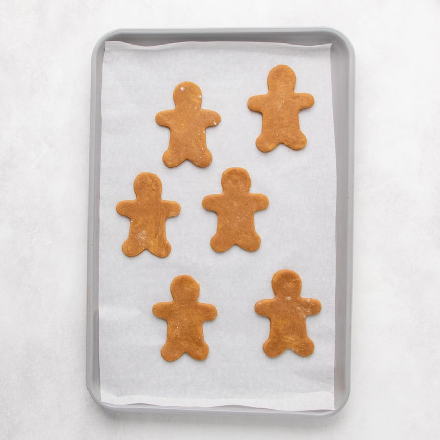 A parchment-lined baking sheet with six unbaked brown gingerbread-person cookies arranged in two columns.