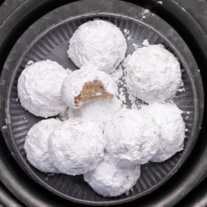 Closeup of a plate piled with powdered-sugar-coated cookies, one showing a bite taken out showing flecks of chopped nuts, all arranged on stacked round metal plates and chargers.