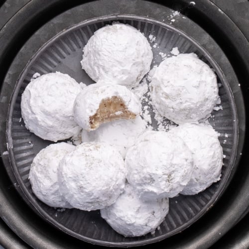 Closeup of a plate piled with powdered-sugar-coated cookies, one showing a bite taken out showing flecks of chopped nuts, all arranged on stacked round metal plates and chargers.