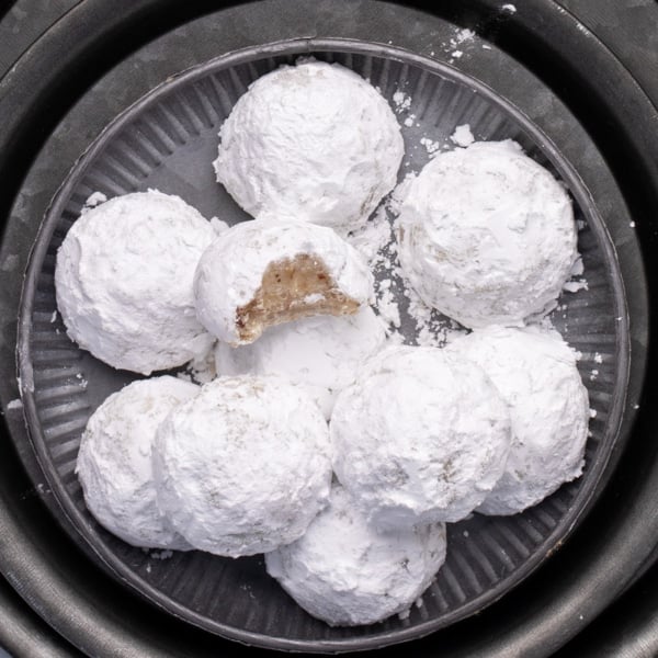 Closeup of a plate piled with powdered-sugar-coated cookies, one showing a bite taken out showing flecks of chopped nuts, all arranged on stacked round metal plates and chargers.