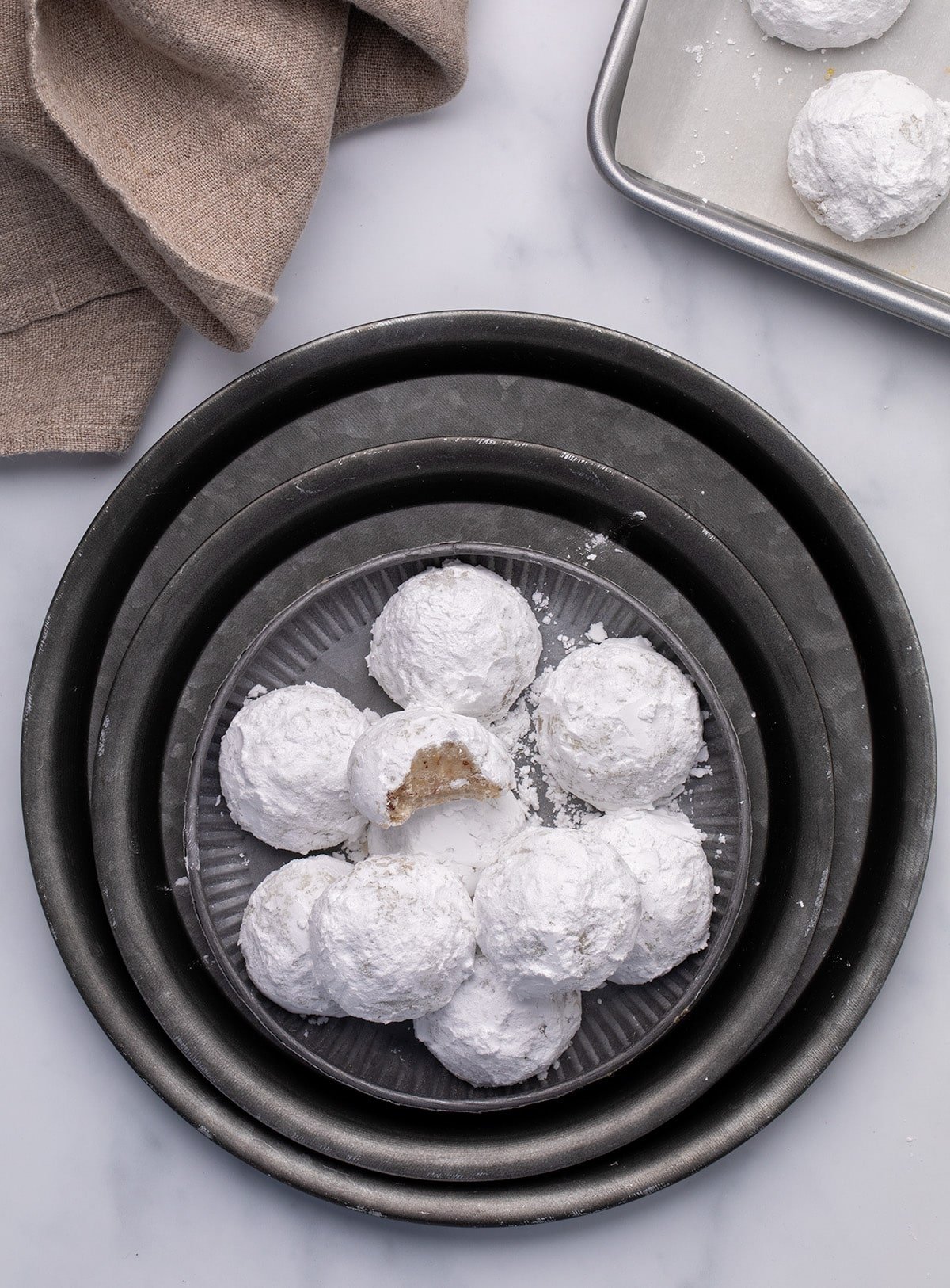 A wider view of powdered-sugar-coated gluten free Mexican wedding cookies piled on a plate set inside stacked metal trays, with a beige cloth and part of a baking sheet visible in the background.