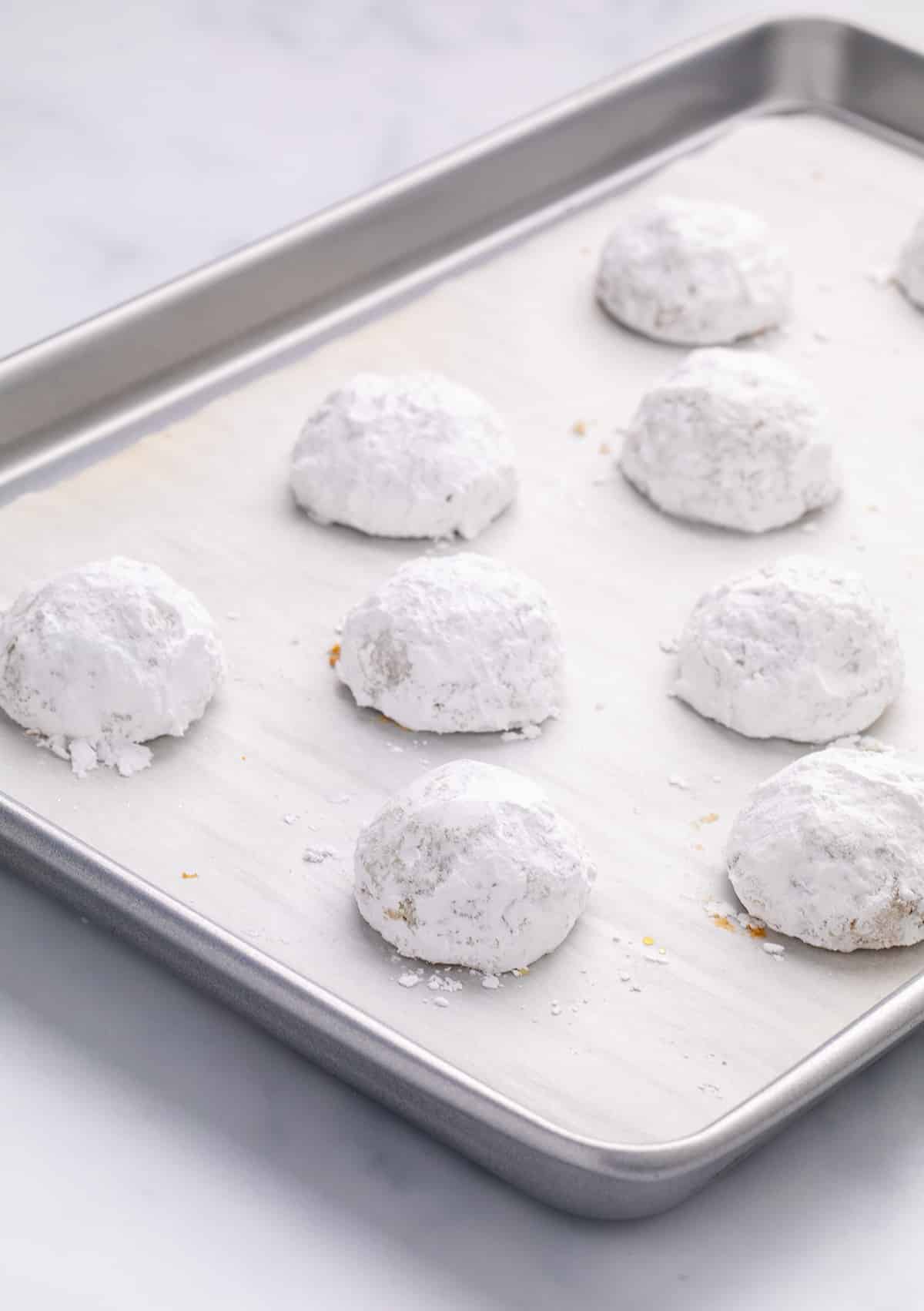 A close view of powdered-sugar-coated cookies on a parchment-lined baking sheet, with the cookies arranged in rows.