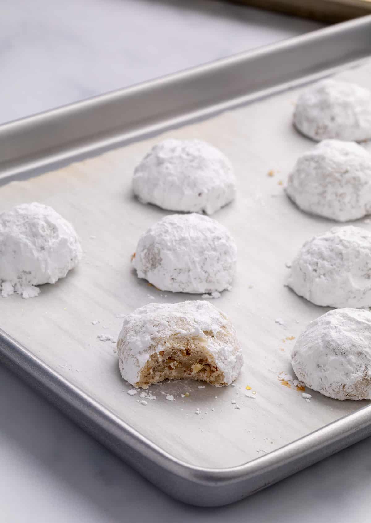 A close view of powdered-sugar-coated cookies on a parchment-lined baking sheet, with one cookie in front showing a bite taken out revealing flecks of chopped pecans inside.
