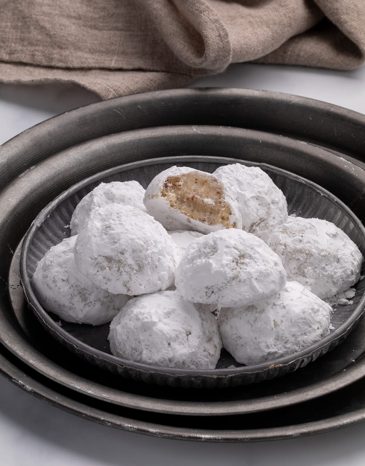 A side angle view of a plate of powdered-sugar-coated cookies stacked on nested metal trays, with one cookie on top showing a bite taken out and a beige cloth in the background.