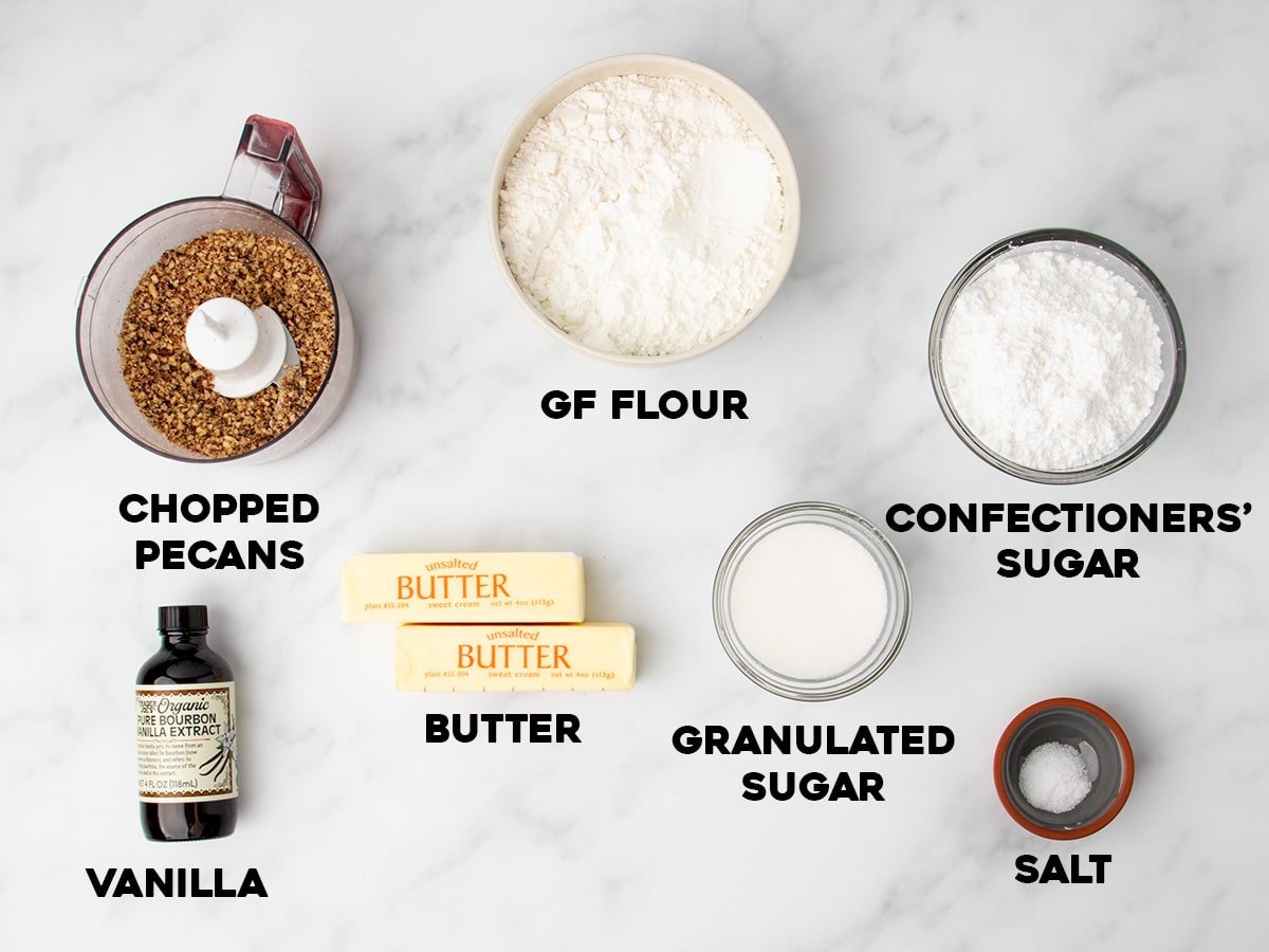 Bowls and containers of ingredients arranged on a marble surface, including chopped pecans, gluten free flour, confectioners’ sugar, vanilla extract, two sticks of butter, granulated sugar, and salt.