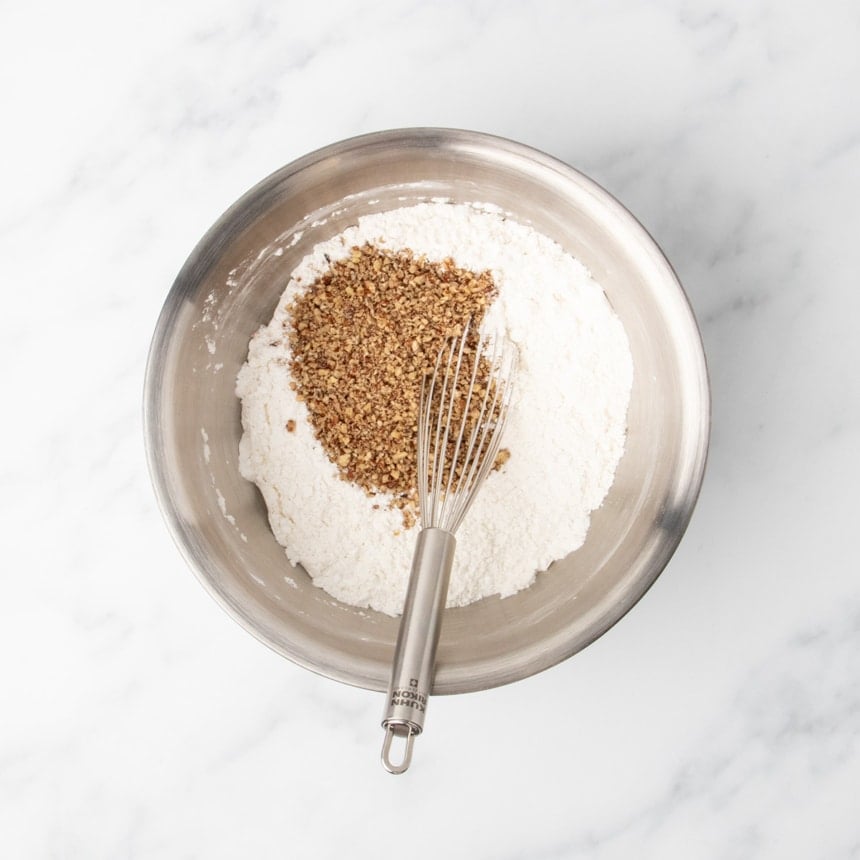 A metal mixing bowl filled with white dry ingredients with a pile of finely chopped nuts on top, and a metal whisk resting inside the bowl.