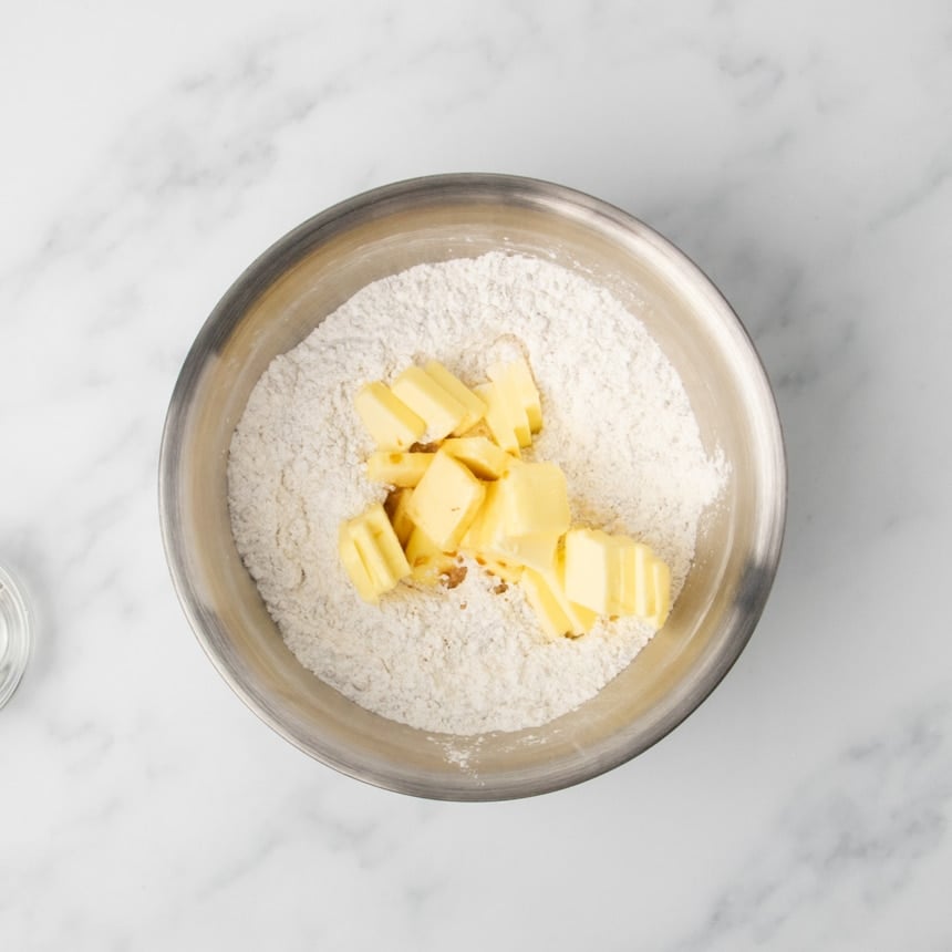 A metal mixing bowl with dry ingredients and several cubes of butter scattered on top.