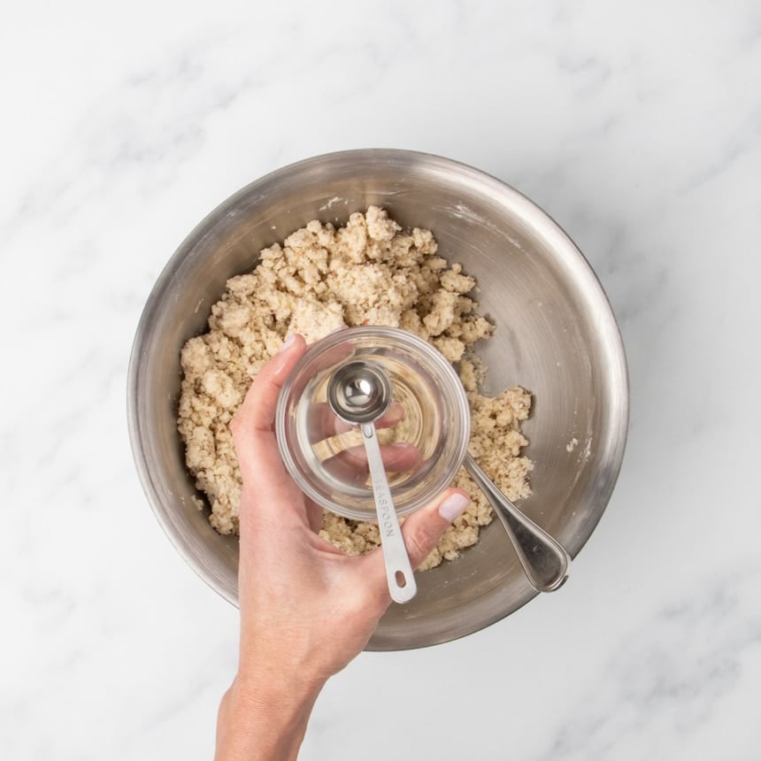 A hand holding a small glass bowl with a metal measuring spoon inside it over a larger bowl of clumps of light tan raw cookie dough.