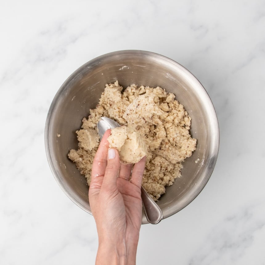 A hand holding a small packed piece of light tan dough with brown flecks taken from a bowl of mixed clumps of raw dough, with a spoon resting inside the bowl.