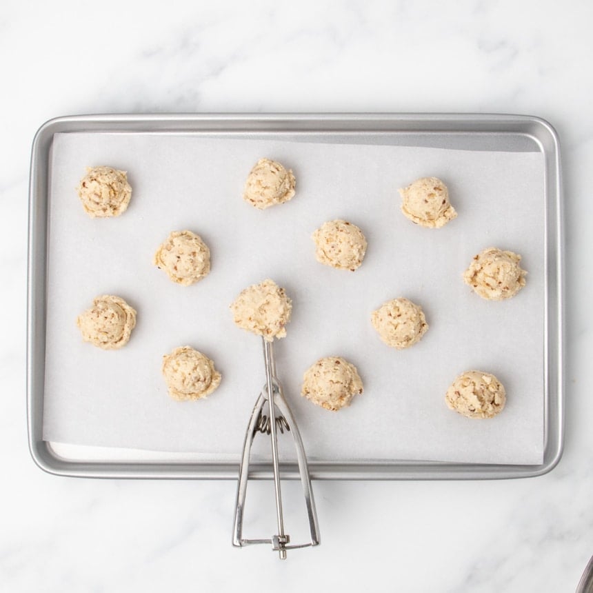 Small scoops of raw light tan cookie dough spaced on a parchment-lined baking sheet, with a metal cookie scoop with more raw dough placed among them.