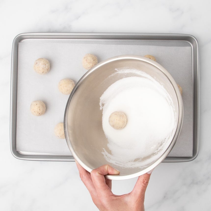 A hand holding a metal bowl partially filled with granulated sugar, with one dough ball sitting in the sugar and other dough balls on a baking sheet below.