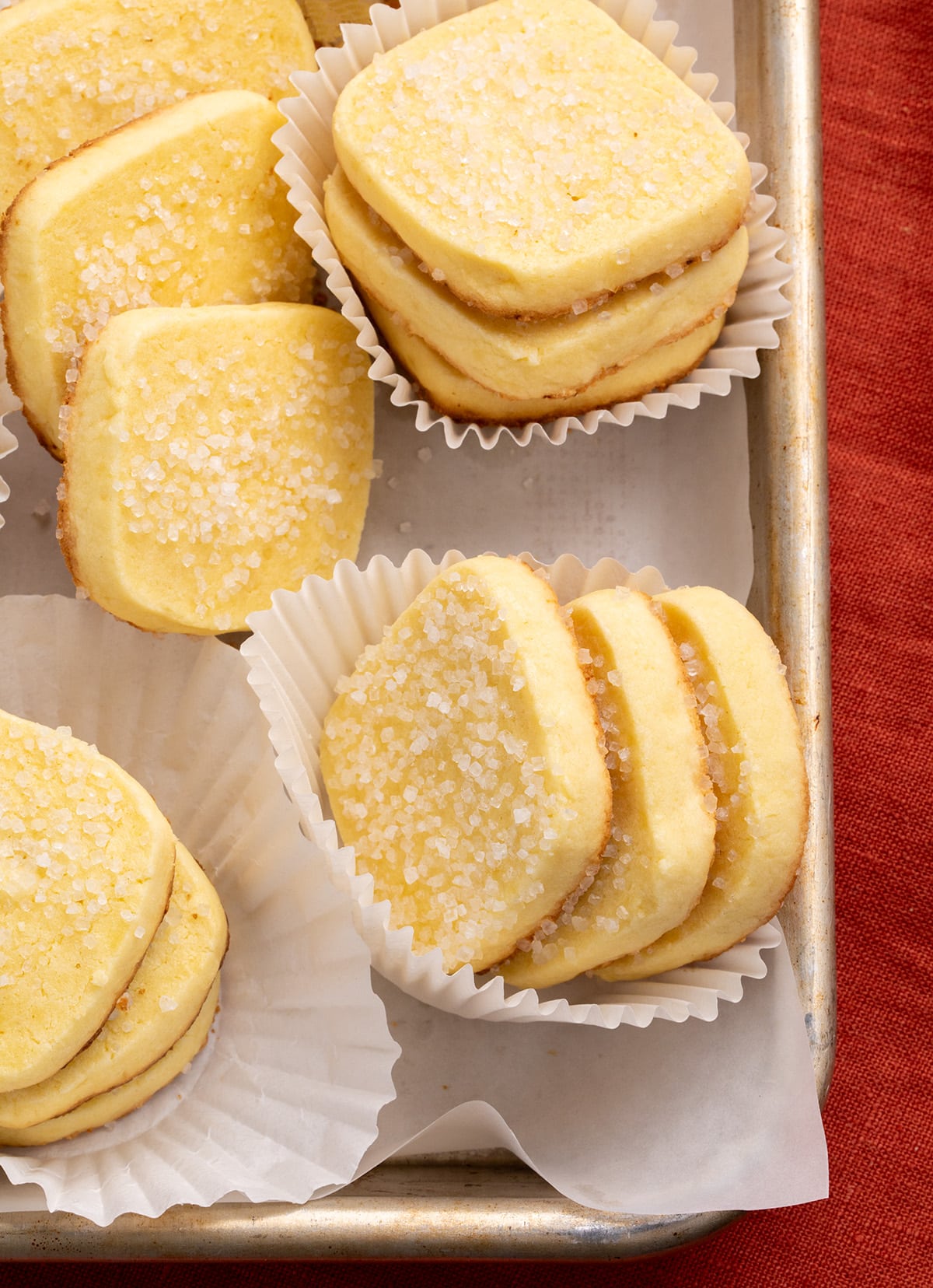 Multiple stacks of yellow rectangular sugar-topped gluten free butter cookies in white paper liners on a metal tray on top of a red cloth.