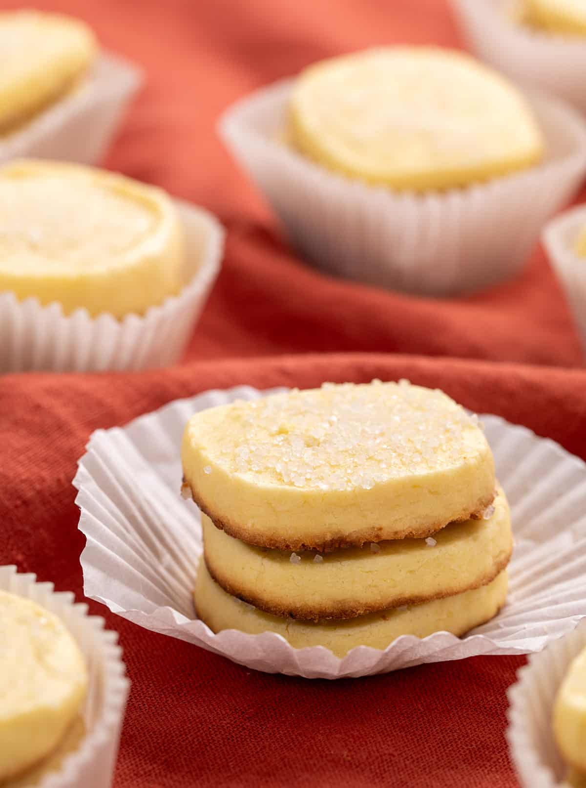 A small stack of baked yellow rectangular cookies with browned bottoms in a white paper liner, surrounded by other cookies on a red textured fabric.