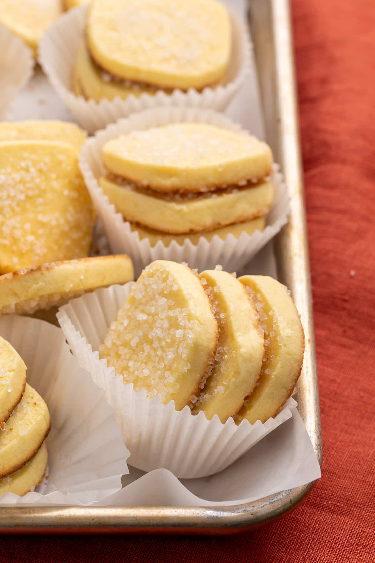 Several stacks of baked rectangular cookies in white paper liners arranged on a metal tray next on top of a red cloth with the lead 3 cookies on their sides showing their browned edges.