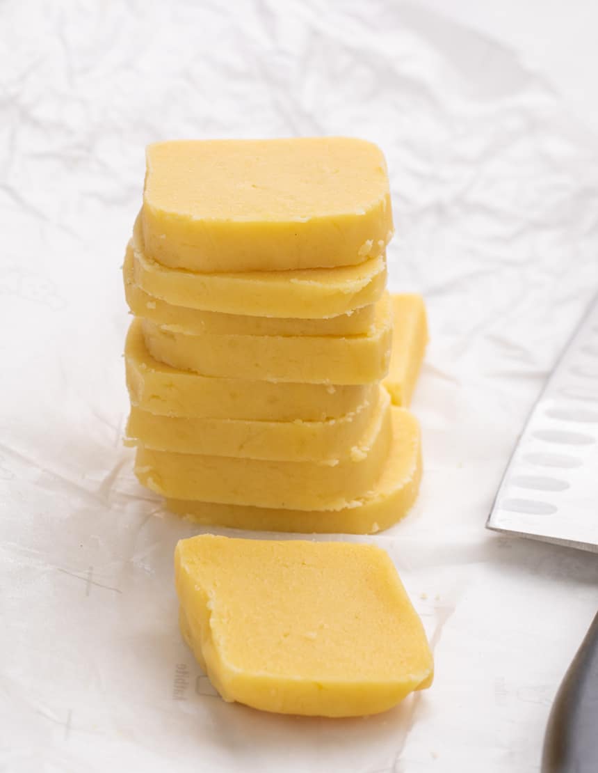 A stack of uncooked rectangular yellow cookie dough slices on parchment paper with an additional slice lying flat beside a knife.