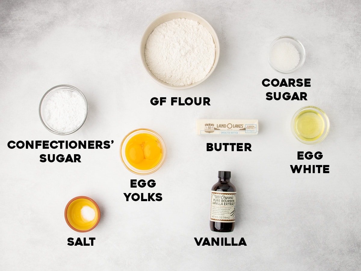 A top view of bowls and containers holding gluten free flour, coarse sugar, confectioners’ sugar, egg yolks, egg white, salt, butter stick, and a bottle of vanilla arranged on a light surface, each ingredient labeled with text.