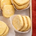 Close-up of several rectangular baked yellow cookies coated with coarse sugar, stacked in white paper liners on a metal tray.