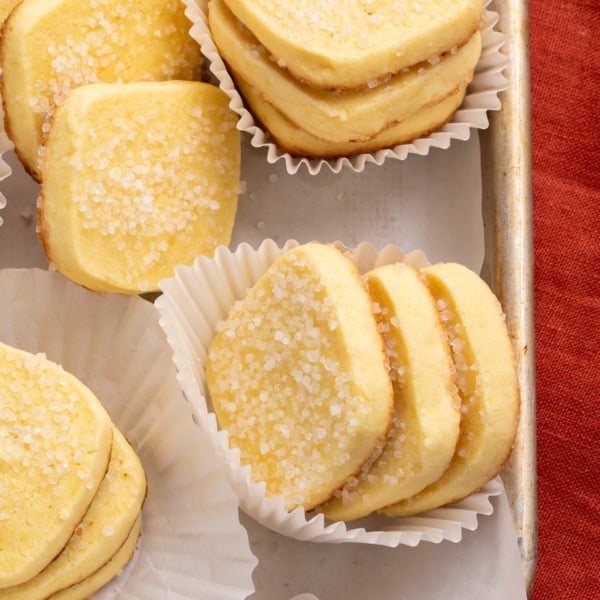 Close-up of several rectangular baked yellow cookies coated with coarse sugar, stacked in white paper liners on a metal tray.