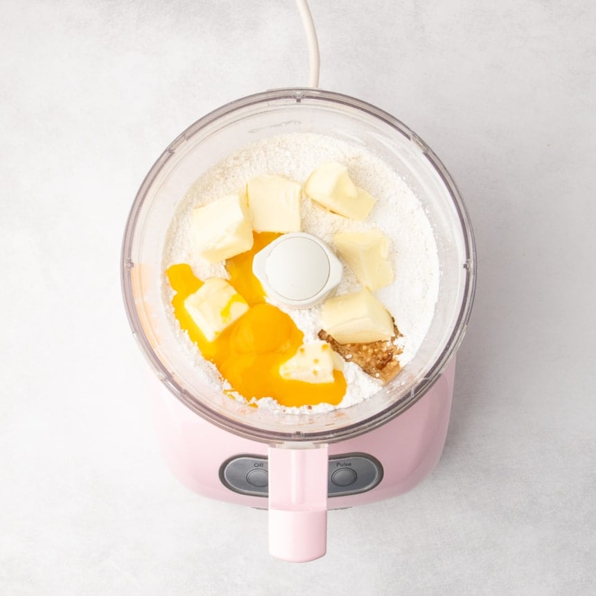 A top view of a food processor bowl filled with white dry ingredients, chunks of butter, egg yolks, and a small amount of vanilla extract.