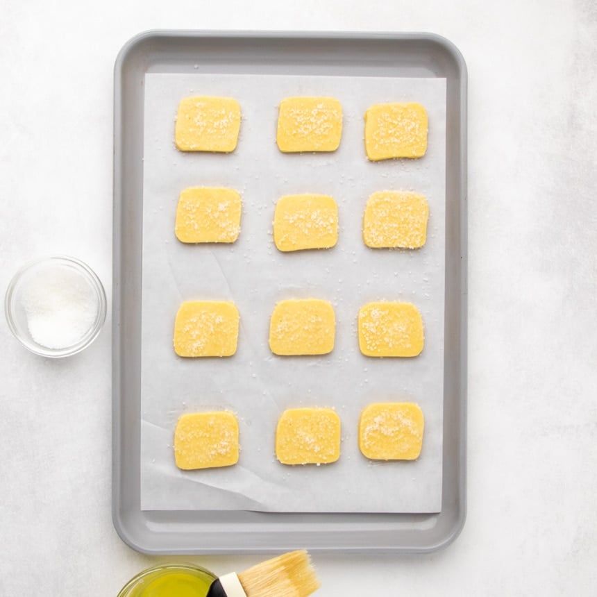 Twelve sugar-topped raw dough pieces arranged in rows on a parchment-lined baking sheet, with a small bowl of sugar visible at the side.