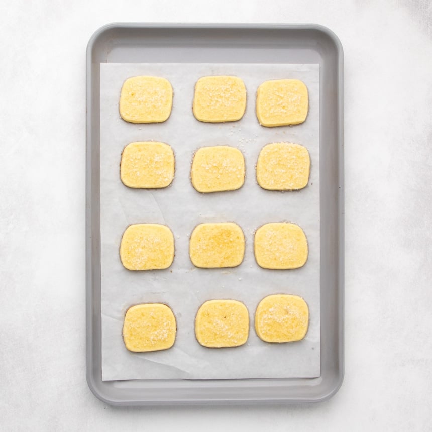 Twelve baked small rectangular yellow cookies with coarse white sugar grains baked into the top, arranged in rows on a parchment-lined baking sheet.