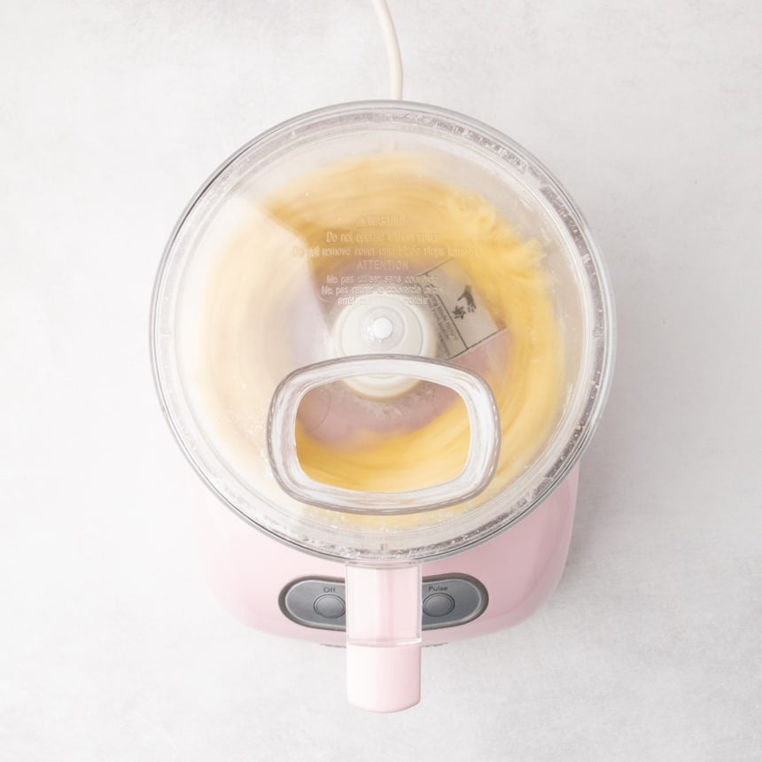 A food processor with the lid on, seen from above, blending a pale yellow mixture in motion.