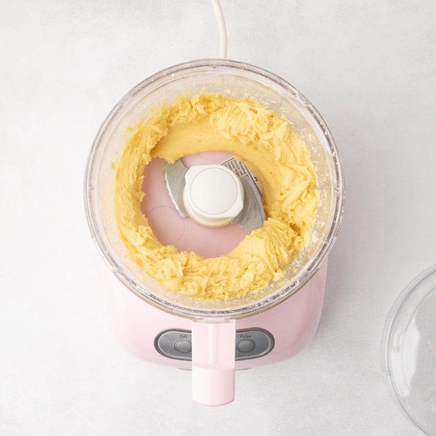 A top view of a food processor bowl containing a smooth, pale yellow dough clinging to the sides in a ring around the central blade.