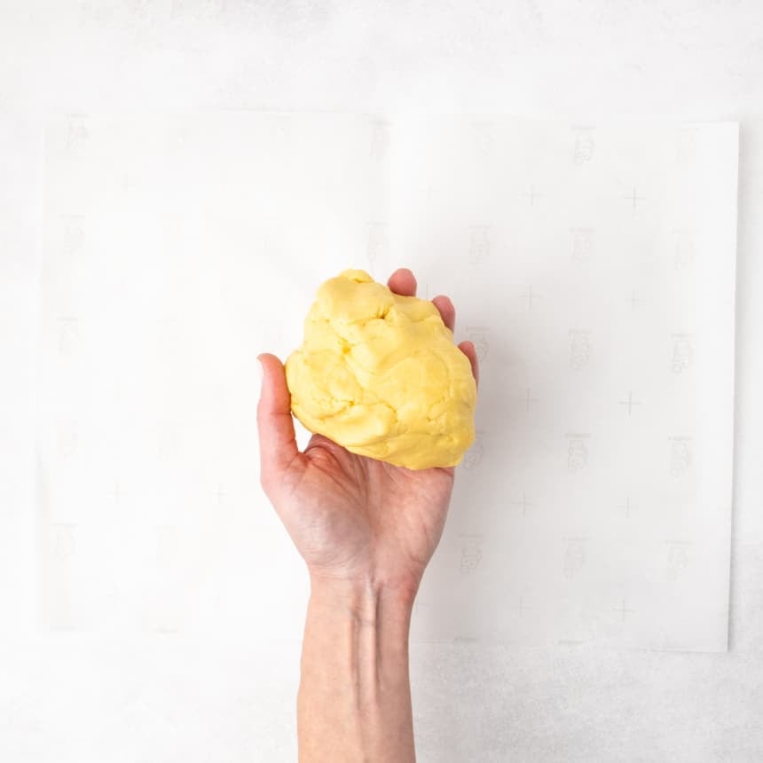 A hand holding a rounded piece of yellow dough over a sheet of parchment paper.