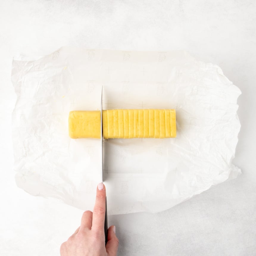 A hand holding a knife and slicing the dough log into even pieces on parchment paper.